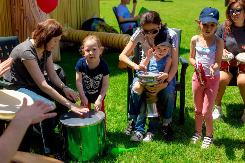 Дети и взрослые играют на музыкальных инструментах на зеленом газоне, создавая веселую атмосферу праздника